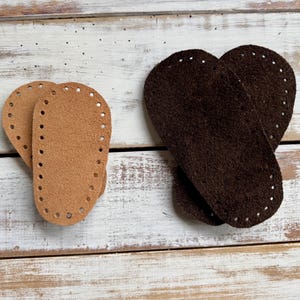 May include: Two pairs of leather soles for crafting projects. One pair is light brown, the other is dark brown. Each sole has evenly spaced holes around the perimeter for stitching. The soles are oval-shaped and appear to be for shoes or bags.