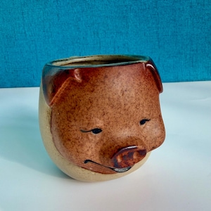 Może przedstawiać: Okrągły ceramiczny kubek w kształcie głowy świni. Kubek ma nakrapianą brązową twarz z wyrzeźbionymi uszami, zamkniętymi oczami i wyraźnym ryjkiem. Dolna część kubka oraz wnętrze są gładkie, w kolorze jasnego beżu z błyszczącym wykończeniem.