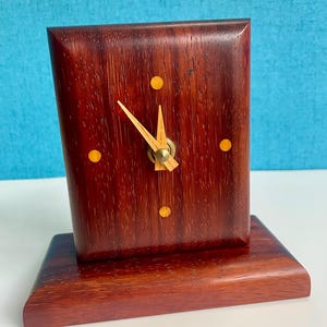 May include: A rectangular wooden desk clock with a dark reddish-brown finish. The clock face has a light-colored wood grain, with three small yellow dots and gold-colored clock hands. The clock sits on a matching rectangular base.