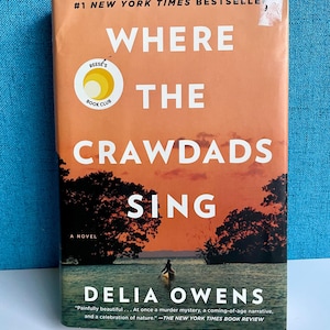 May include: A paperback book cover featuring a sunset scene and the title "Where the Crawdads Sing" by Delia Owens. The book is a #1 New York Times bestseller and a Reese's Book Club pick.