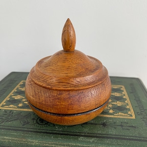 May include: A small, round wooden box with a pointed lid. The box is made of light brown wood and has a dark brown band around the edge.
