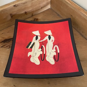 Puede incluir: Bandeja decorativa cuadrada con borde negro e interior rojo. La bandeja presenta un gráfico de dos figuras con ropa tradicional blanca y sombreros cónicos, montando en bicicleta. Las figuras están sobre un fondo rojo.