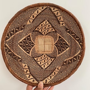 May include: A round, woven basket with a detailed geometric pattern in shades of brown and tan. The basket has a woven rim and a central square design. The basket is held by a hand.