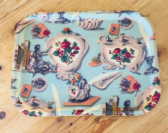 Puede incluir: Bandeja rectangular con fondo azul claro y un patrón repetido de arreglos florales, estatuas y libros. La bandeja tiene un borde redondeado y un acabado brillante. Los arreglos florales presentan rosas rojas y rosas.