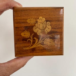 May include: A small, square wooden box with a floral inlay design. The box is a warm brown color with a lighter wood flower and leaf pattern on the lid. The box is held in a person's hand.