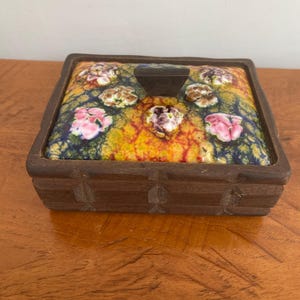 May include: A rectangular wooden box with a dark brown finish and carved side details. The lid features a vibrant, multi-colored ceramic inlay with a crackled texture, decorated with raised floral motifs in pink, white, and purple hues.