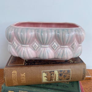 May include: A rectangular, pink ceramic planter with a scalloped design and diamond accents. The planter is resting on a stack of vintage books with brown, green, and red covers.