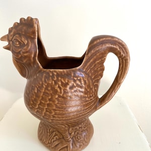 Puede incluir: Jarra de cerámica marrón con forma de gallo y diseño texturizado detallado. La jarra tiene forma de gallo con asa y pico. El gallo tiene una cresta y barbillas detalladas. Es un artículo decorativo.