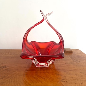 Puede incluir: Una cesta de cristal de color rojo vibrante con una base cuadrada de bordes ondulados y plegados, y un asa alta y arqueada que se estrecha en la parte superior. El cristal pasa de un rojo intenso y saturado en el centro a transparente.