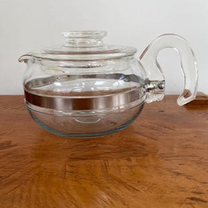 May include: A clear glass teapot featuring a rounded body, a matching glass lid, and a curved glass handle. A metallic copper-coloured band encircles the centre of the glass vessel. The word Pyrex is embossed on the lid and the glass body.