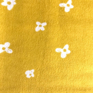 May include: Close-up of a mustard yellow fabric featuring a repeating pattern of white, four-petal flower shapes. The fabric has a soft, textured appearance, with the flowers evenly spaced across the surface.
