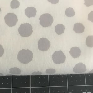 May include: Close-up of a white fabric with a pattern of large, evenly spaced grey polka dots. The fabric is soft and textured. A black ruler with white markings is at the bottom, showing measurements from 66 to 76 cm.