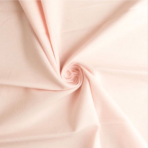 May include: A solid light pink fabric with a smooth, matte texture. The material is gathered into a central swirl, creating soft, radiating folds and shadows that highlight the consistent pastel color and the fine, uniform weave of the textile.