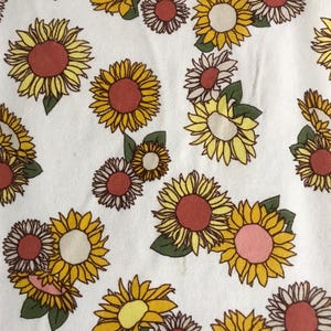 Op de afbeelding: Witte stof met een herhaald patroon van zonnebloemen in geel, oranje en roze. De zonnebloemen hebben groene bladeren en zijn verspreid gerangschikt.