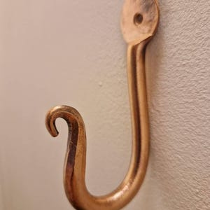 May include: A copper-colored, J-shaped wall hook. The hook has a rounded top with a single hole for mounting and a curled end. The metal has a slightly aged appearance.