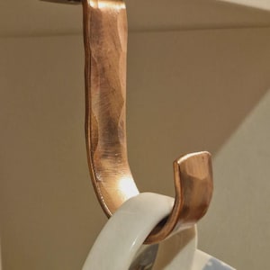 May include: A copper mug hook with a textured finish. The hook is mounted on a white surface and holds a white and blue ceramic mug. The hook is designed to be mounted on a wall or cabinet.
