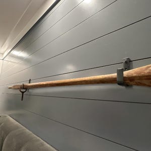 May include: A vintage wooden oar mounted on a gray shiplap wall with metal brackets. The oar is a light brown color and appears to be aged. The wall is painted a solid gray color. The oar is a decorative nautical element.