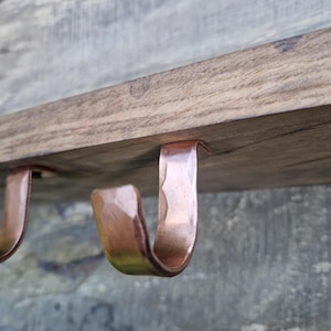 May include: Close-up of a wooden shelf with three copper-coloured, hammered metal hooks attached to the underside. The wood has a natural grain and a warm brown tone. The hooks are curved and have a textured surface.