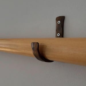 May include: A light-coloured wooden rod mounted horizontally with a dark brown, textured metal bracket. The bracket features a curved hook shape and is secured to the wall with two silver-coloured Phillips head screws visible on the mounting plate.