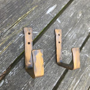 May include: Two L-shaped wall hooks made of hammered metal with a bronze-toned finish. Each hook features a flat vertical mounting plate with two pre-drilled circular holes for installation and a curved upward-facing arm for hanging items.