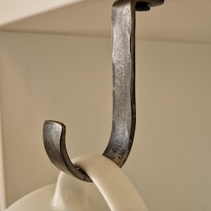 May include: Hand-forged iron mug hook, designed to hang from a shelf. The hook has a textured, dark grey finish and a curved lower section to hold a mug. A white mug handle is visible.