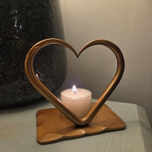 May include: A gold-coloured metal candle holder shaped like a heart, with a small lit candle inside. The heart frame sits on a matching rectangular base. The background includes a dark blue vase.