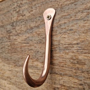 May include: A copper-coloured metal hook fixed to a wooden surface. The J-shaped hook has a curved end and is secured with a single screw. The wood displays a natural grain pattern.