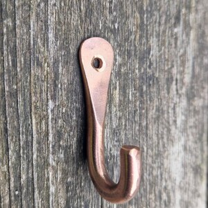 May include: A copper-coloured metal hook with a curved design. The hook has a rounded end and a hole at the top for mounting. The hook is shown against a weathered wooden background.