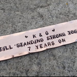 May include: A copper-coloured metal bar with the text "K & G STILL STANDING STRONG TOGETHER 7 YEARS ON" stamped on it. The bar has a hammered texture and rounded edges. Small heart symbols are stamped above the letters "K & G".