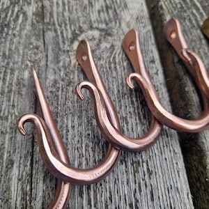 May include: Close-up of several copper-colored metal hooks. The hooks have a curved design with a pointed end and a looped hook. The hooks are mounted on a wooden surface, showcasing their rustic aesthetic. These hooks are ideal for hanging items.