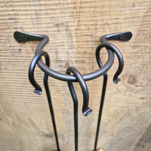 May include: Black wrought iron fireplace tool set with a shovel, poker, and brush. The set is mounted on a wooden background. The holder has a unique, curved design.