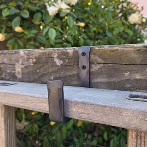 May include: Close-up of a weathered wooden ladder with metal brackets and hooks. The ladder is grey and brown, with visible wood grain. The metal components are dark grey. The background features green foliage and white flowers.