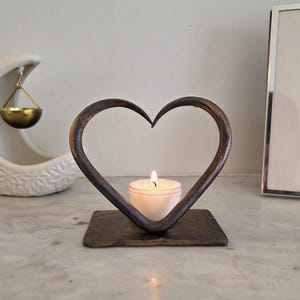 May include: A dark brown, hand-forged metal candle holder shaped like an open heart, featuring a flat rectangular base and a curved frame that holds a single small white tea light candle with a lit flame in the center of the heart shape.