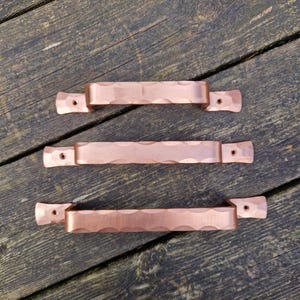 May include: Three copper-coloured cabinet pulls with a textured, wavy design. Each pull has a rectangular bar with curved indentations and a flat end with a single drilled hole for mounting. The pulls are displayed on a weathered wooden surface.