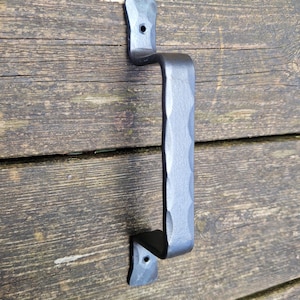May include: A black, hammered metal door pull handle. The handle is mounted on a weathered wooden surface. The handle has a textured, dimpled grip and is attached with two mounting plates, each with a single hole.