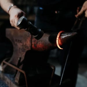 May include: A blacksmith forges a metal ring on an anvil. The ring is glowing red-orange from the heat. The blacksmith is using a hammer and tongs to shape the metal. The anvil is dark and weathered.