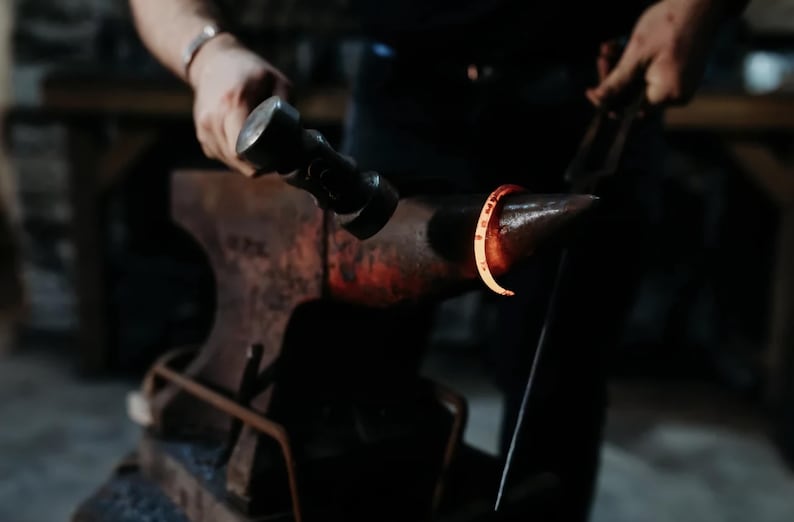 May include: A blacksmith forges a metal ring on an anvil. The ring is glowing red-orange from the heat. The blacksmith is using a hammer and tongs to shape the metal. The anvil is dark and weathered.