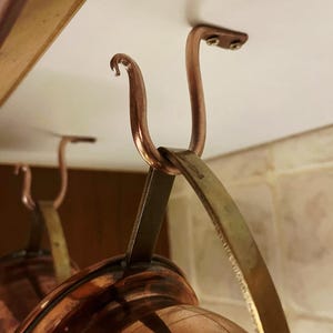 May include: Close-up of a copper pot rack with curved hooks and a brass support. The rack is mounted on a white ceiling, with the copper pots hanging below. The copper has a shiny, reflective surface.
