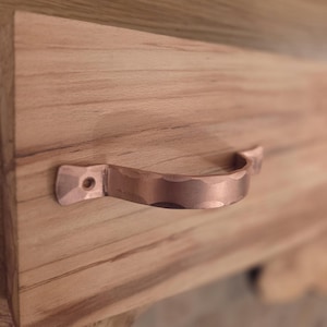 May include: A copper-coloured drawer pull with a textured, curved handle is attached to a light-coloured wooden surface. The handle has a hammered appearance and is mounted with two small screws.