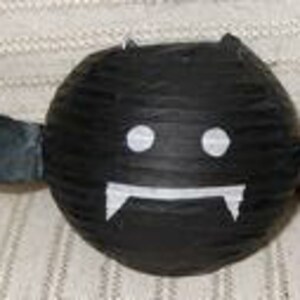 May include: A handmade, round, black bat decoration with white eyes and fangs. The bat has two small horns and two black wings extending from its sides. The bat is made of paper or cardboard.