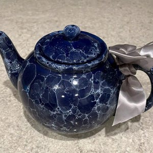 May include: A navy blue ceramic teapot with a marbled white and blue pattern. The teapot has a curved spout, a rounded body, and a lid with a small knob. A silver ribbon is tied around the handle.