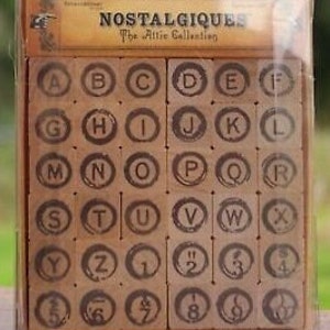 May include: A set of 36 wooden alphabet stamps in a clear plastic case. The stamps are circular and feature a vintage-inspired design. The letters are in uppercase and include numbers 1-9.