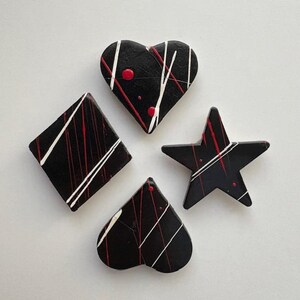 May include: Four black decorative shapes: a heart, diamond, star, and spade. Each shape has white and red paint streaks and splatters. The shapes are arranged on a white surface.