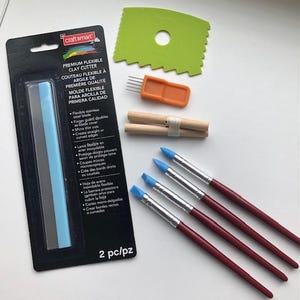 May include: A collection of clay sculpting tools, including a flexible clay cutter, brushes with blue tips, and other shaping tools. The tools are arranged on a white surface, with a black and gray package of the clay cutter visible.