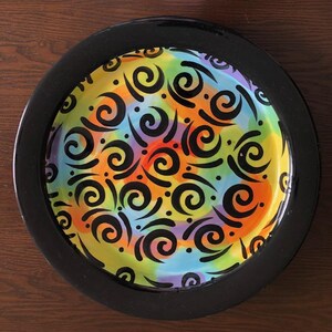 May include: A round ceramic plate with a black rim. The plate's interior features a vibrant, colorful design with swirls and dots in orange, yellow, green, purple, and red. Black spiral patterns are layered over the colorful background.
