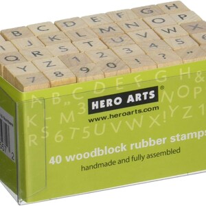 May include: A clear plastic box containing 40 wooden block rubber stamps with letters, numbers, and punctuation marks. The box is green and has the text "HERO ARTS" and "www.heroarts.com" printed on it. The text "40 woodblock rubber stamps" and "handmade and fully assembled" is also printed on the box.