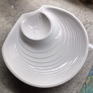 May include: White ceramic serving dish shaped like a seashell with two separate bowls. The dish has a textured, rippled surface.
