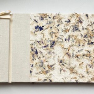 May include: A handmade journal with a cream-colored fabric spine and cover. The right side of the journal features a textured paper embedded with dried blue and tan flower petals. The left side has a cream-colored fabric cover with a ribbon tie.
