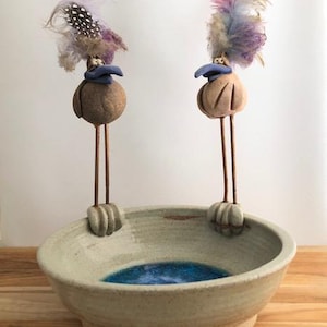 May include: Two ceramic birds with long legs and colorful feathers stand on the edge of a light green ceramic bowl with a blue glaze inside.