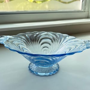 May include: A light blue glass serving bowl with a scalloped rim and a pedestal base. The bowl has a detailed, swirling pattern and two decorative handles. The bowl is approximately 10 inches in diameter.